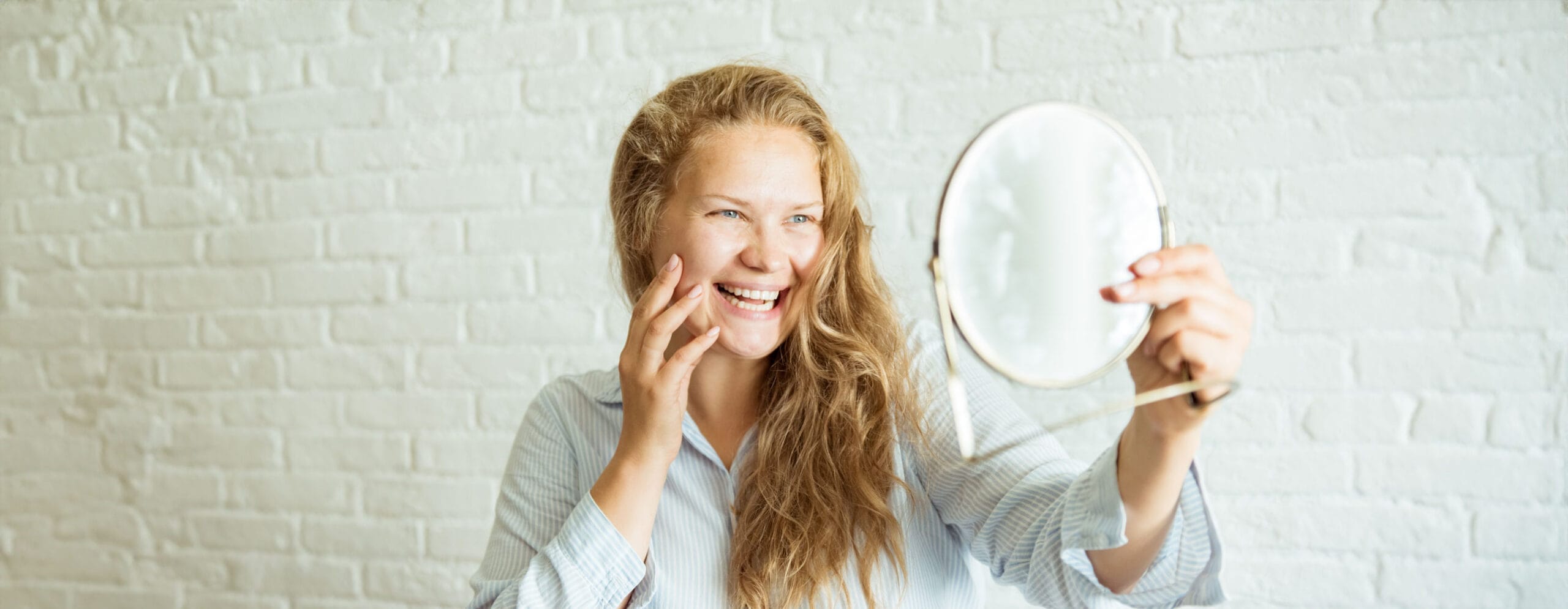 happy-woman-applying-cream-looking-mirror-grey-background-scaled